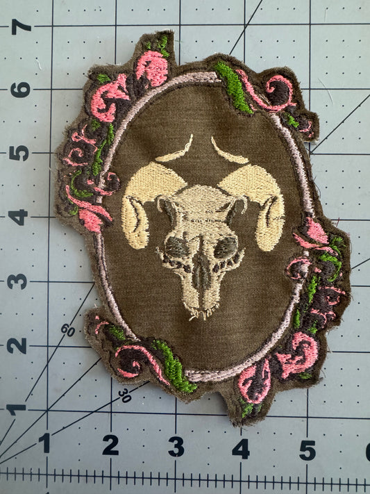 Two embroidered patches featuring a horned ram skull centered within ornate Baroque-style frames—one on brown fabric with pink and green scroll accents, the other on light gray with brighter threads.