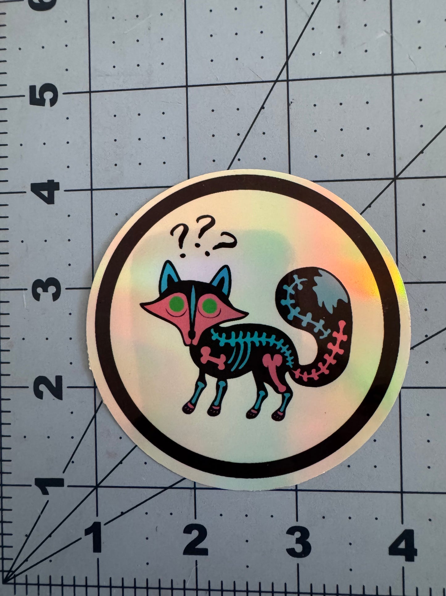 A round holographic sticker featuring a confused neon skeletal fox with a pink face, green eyes, and bright blue and magenta bones. Three question marks float above its head. The fox is outlined in black and centered within a white circle bordered in black.