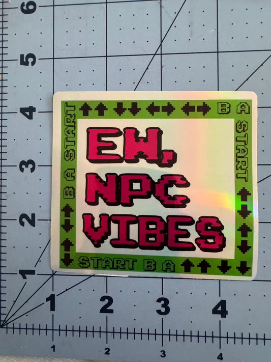 Two versions of a square sticker featuring pixelated text reading “Ew, NPC Vibes” in red on a white background. The border is bright green with black 8-bit arrow symbols and the words “START B A” repeated, referencing a classic video game controller pattern. One version has a standard white finish, the other has a holographic sheen. Stickers are shown on a blue grid cutting mat with inch measurements for scale.