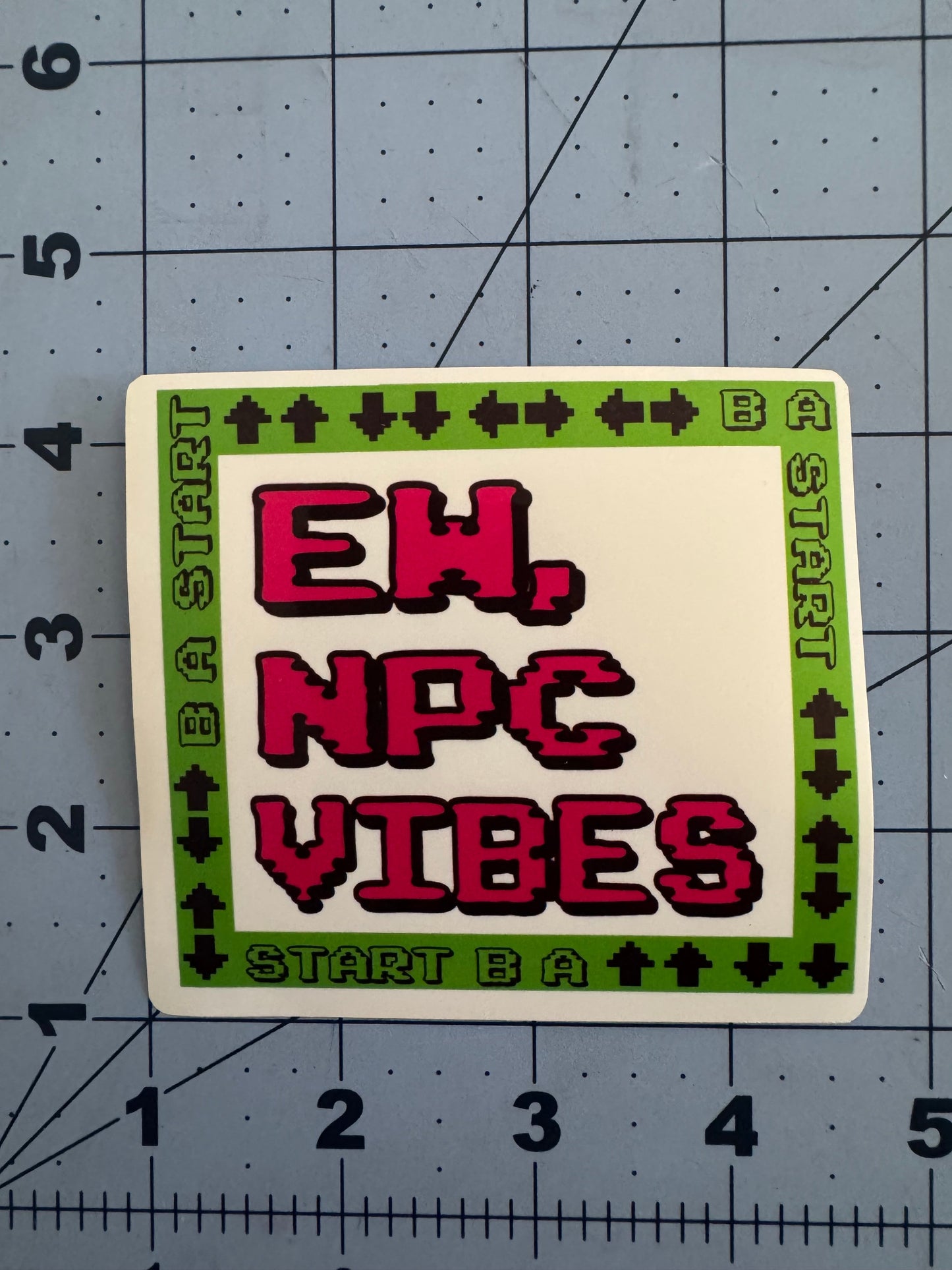 Two versions of a square sticker featuring pixelated text reading “Ew, NPC Vibes” in red on a white background. The border is bright green with black 8-bit arrow symbols and the words “START B A” repeated, referencing a classic video game controller pattern. One version has a standard white finish, the other has a holographic sheen. Stickers are shown on a blue grid cutting mat with inch measurements for scale.