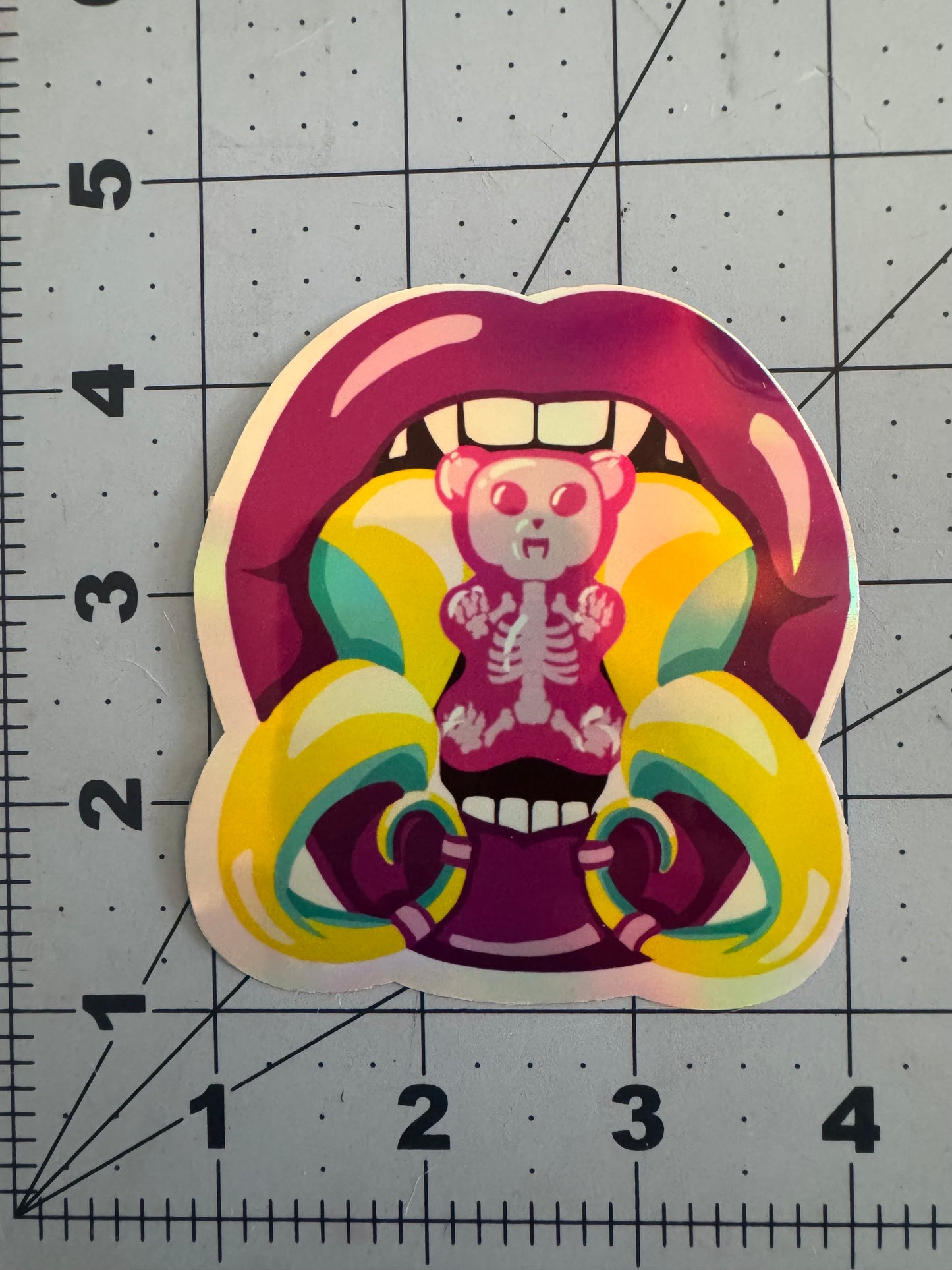 A holographic sticker featuring a pair of glossy magenta lips biting into a rainbow-colored gummy bear with a visible skeleton inside. The bear is centered between two sharp vampire fangs and framed by bright yellow and teal spiral fangs piercing the lower lip, adding a surreal and edgy vibe.
