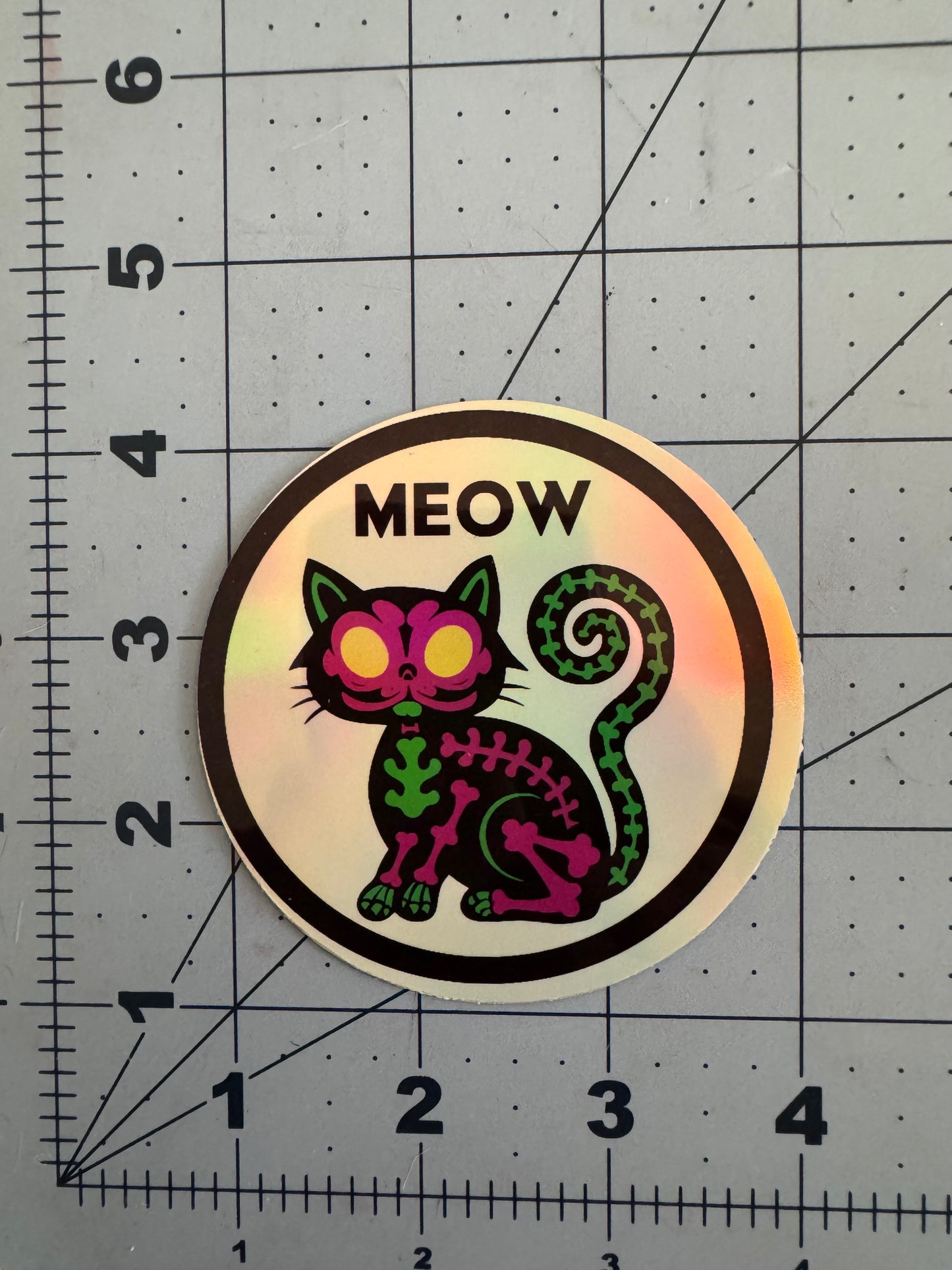 Holographic round sticker featuring a neon skeletal cat with glowing yellow eyes, curled tail, and the word “MEOW” above its head. The cat’s bones are illustrated in green and pink with a bold black border.