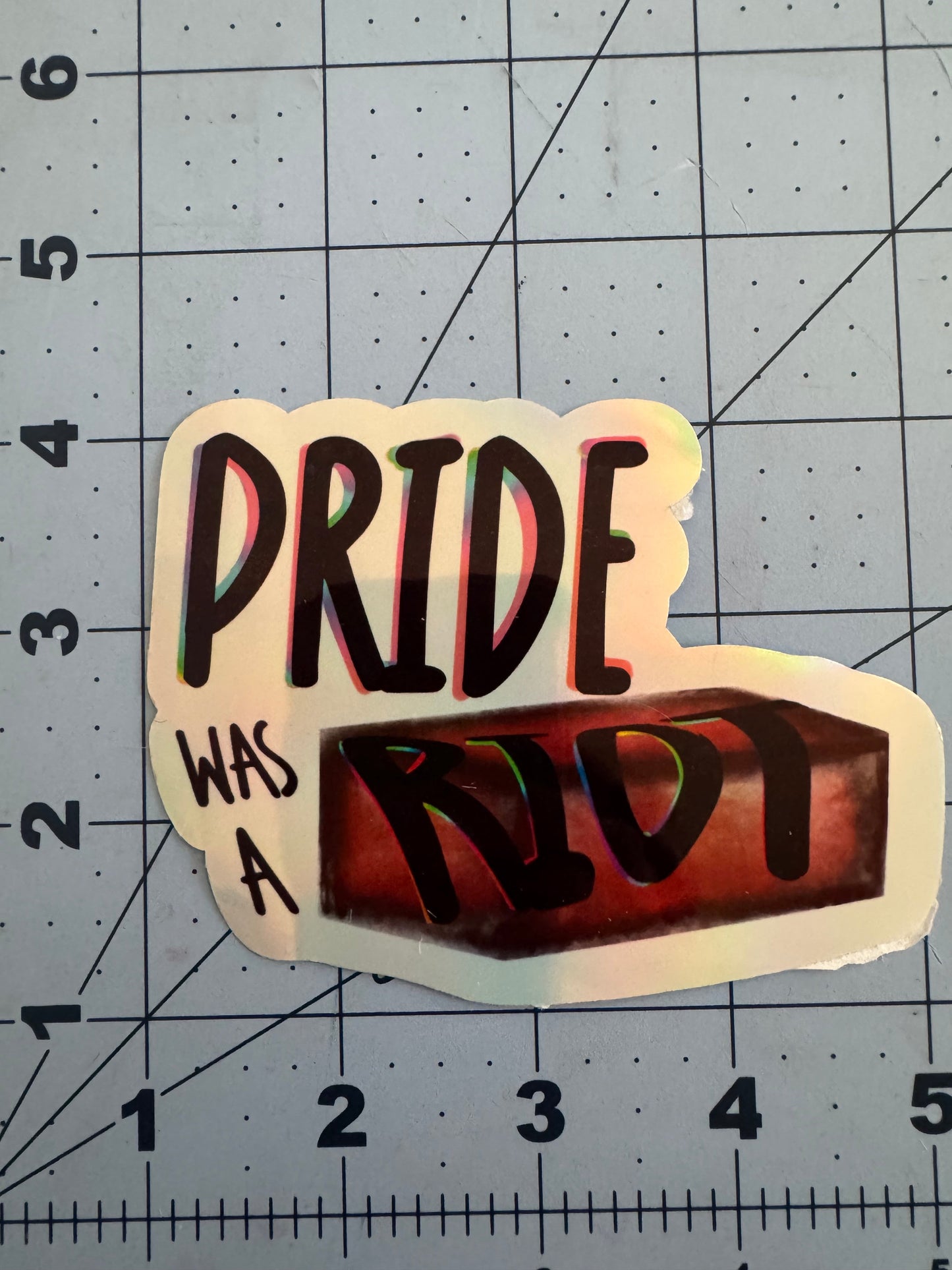 Sticker reading “Pride Was a Riot” with the word “Riot” stylized as a red brick. Available in holographic and classic white finish.