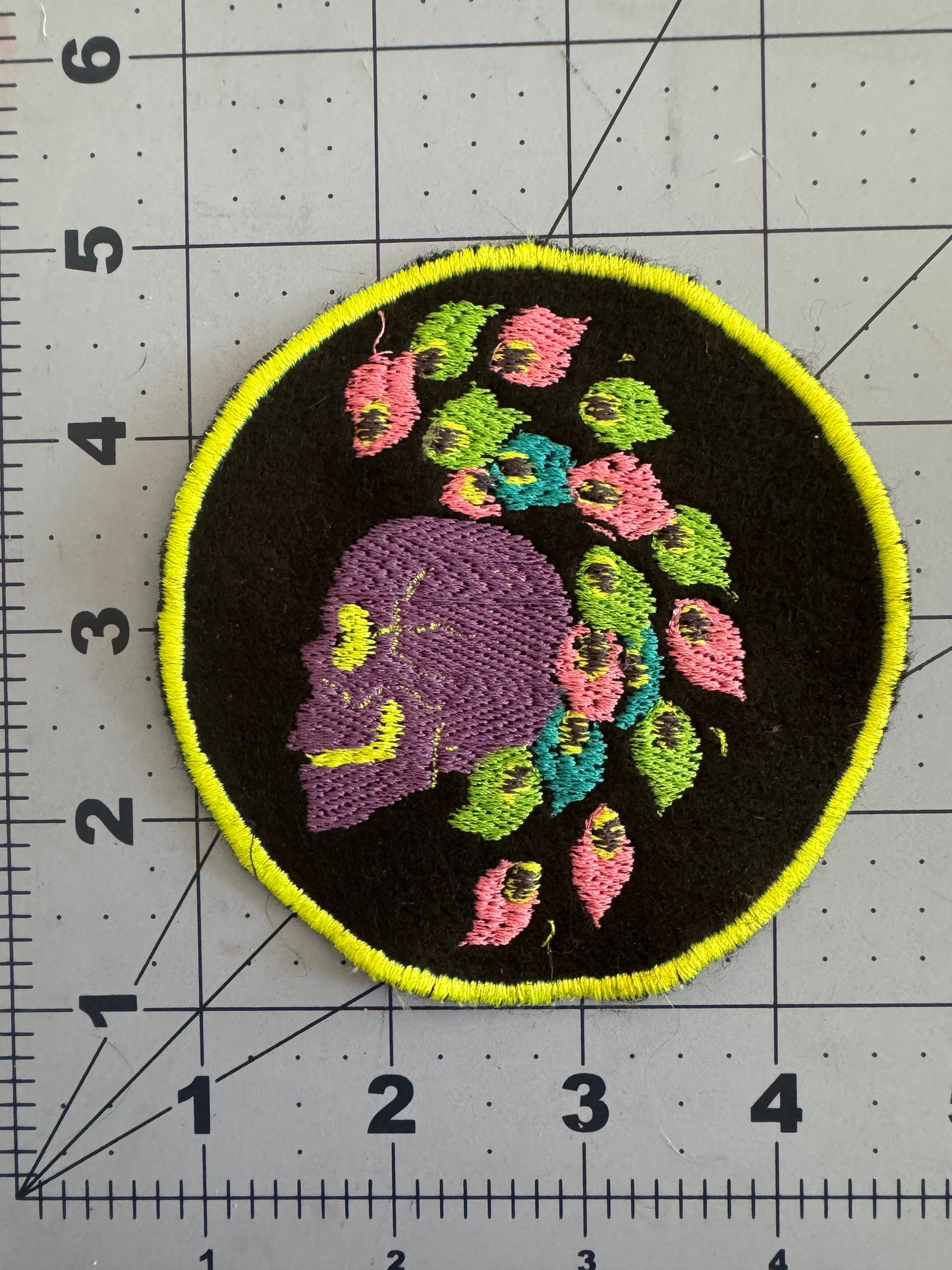 Two embroidered circular patches featuring side-profile skulls with bright mohawk hairstyles made of stylized neon flowers and leaves. One skull is yellow with pink and teal details and a hot pink border; the other is purple with multicolored flowers and a neon yellow border.