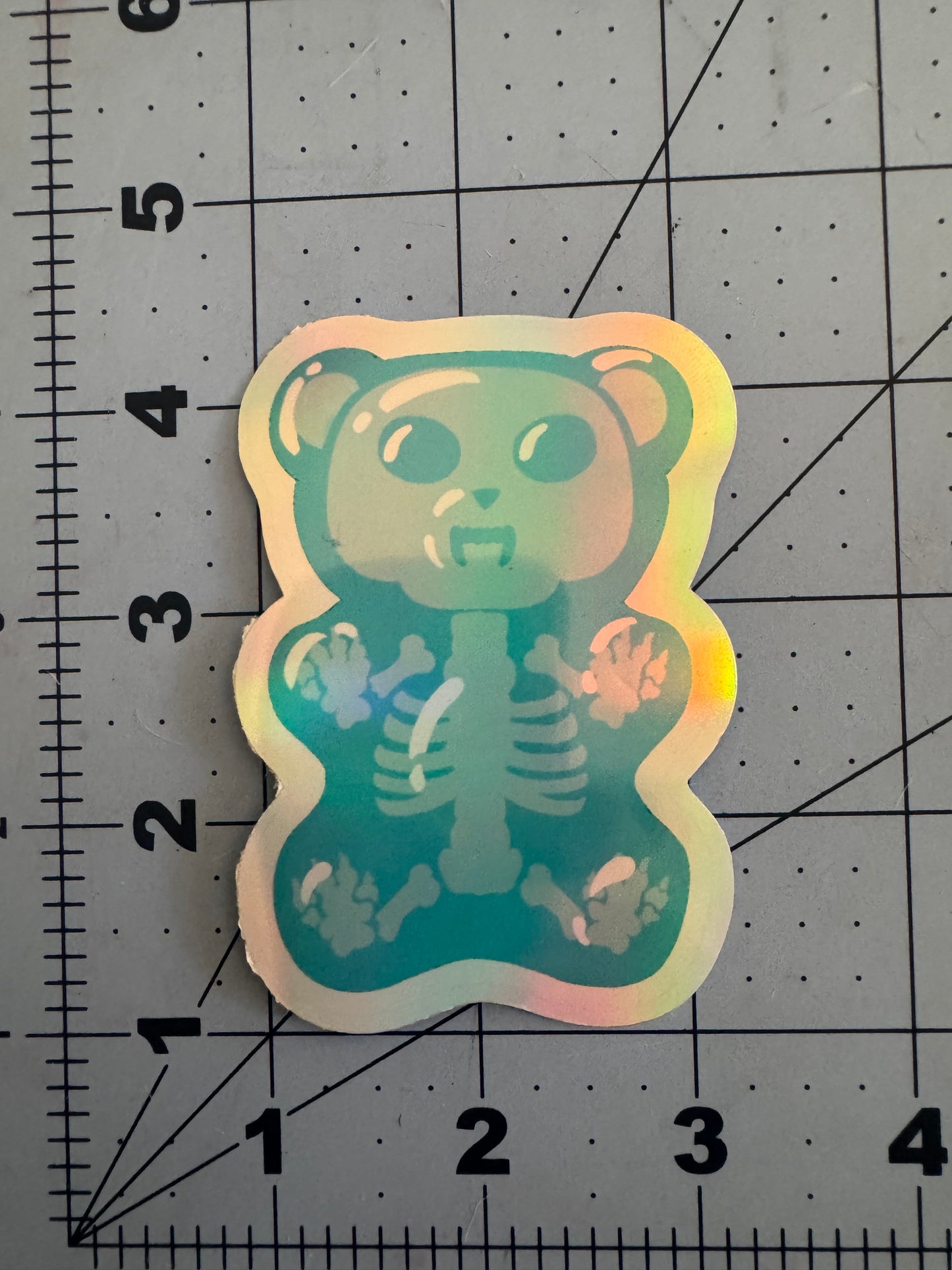 Two holographic vinyl stickers featuring cartoon-style gummy bears with fanged expressions and translucent bodies revealing skeletal structures inside; one is blue, the other green, both displayed on a measurement grid.