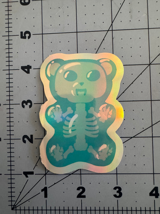 Two holographic vinyl stickers featuring cartoon-style gummy bears with fanged expressions and translucent bodies revealing skeletal structures inside; one is blue, the other green, both displayed on a measurement grid.