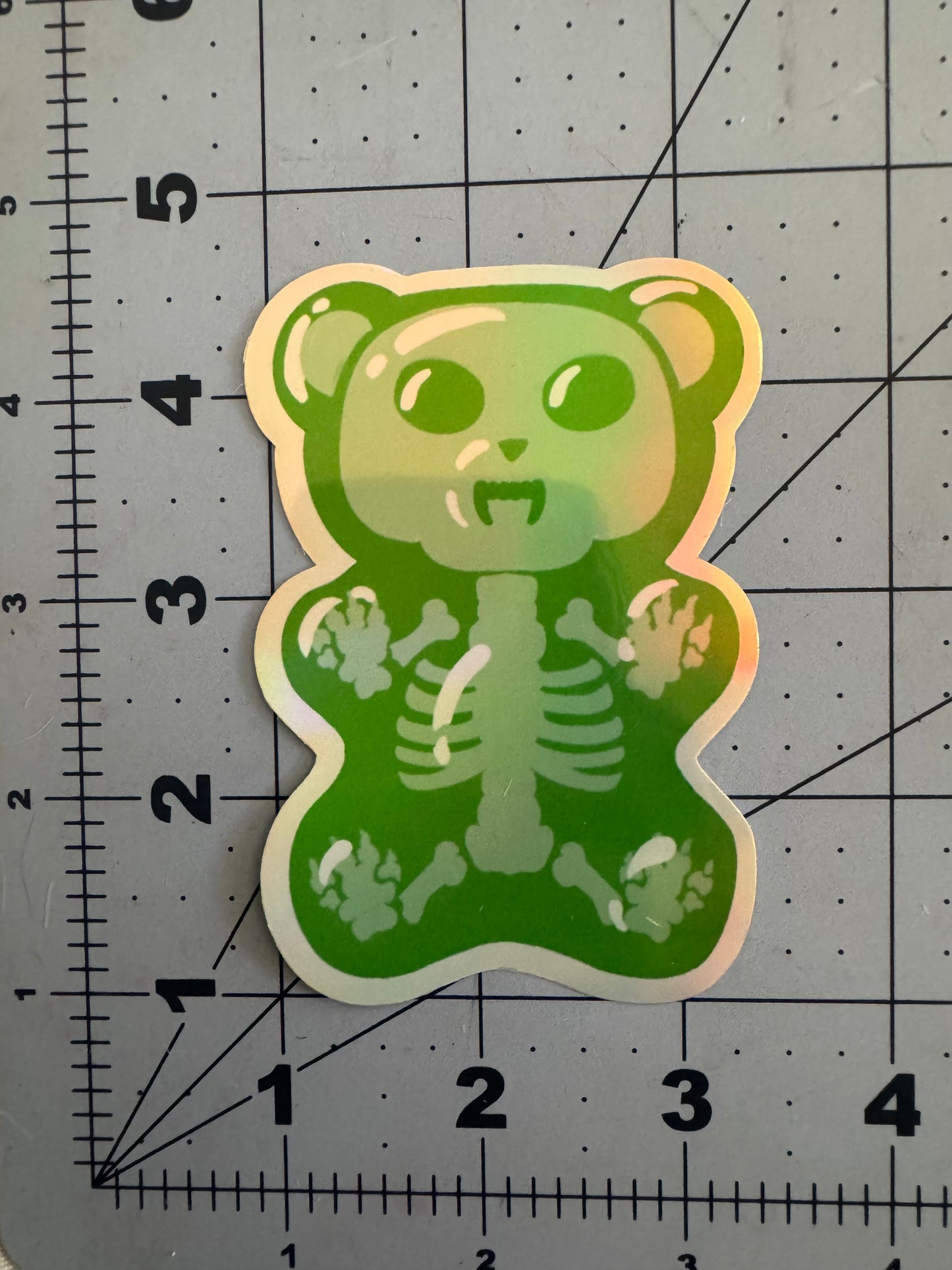 Two holographic vinyl stickers featuring cartoon-style gummy bears with fanged expressions and translucent bodies revealing skeletal structures inside; one is blue, the other green, both displayed on a measurement grid.