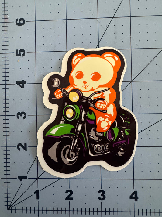 White vinyl sticker of a skeletal gummy bear riding a green motorcycle, with bold black outlines and colorful accents. The sticker is placed on a cutting mat with a measurement grid, showing it is approximately 3 inches tall.