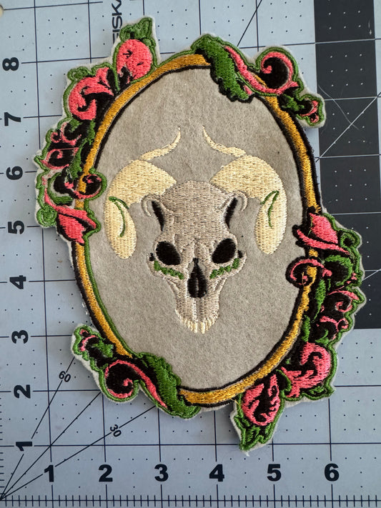 Two embroidered patches featuring a horned ram skull centered within ornate Baroque-style frames—one on brown fabric with pink and green scroll accents, the other on light gray with brighter threads.