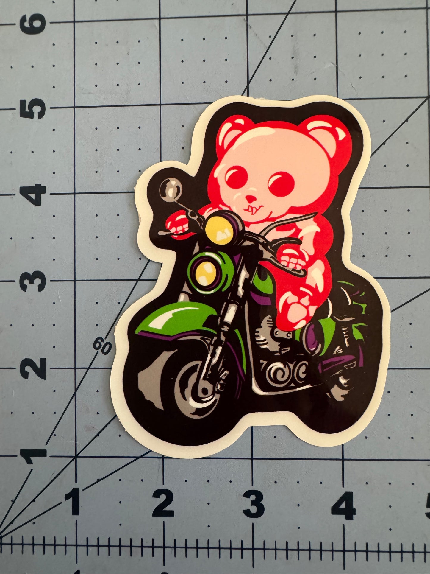 A classic white vinyl sticker featuring a red and white skeletal gummy bear riding a green and purple motorcycle. The bear grips the handlebars with a determined expression. The sticker is placed on a blue grid background with inch measurements for scale.