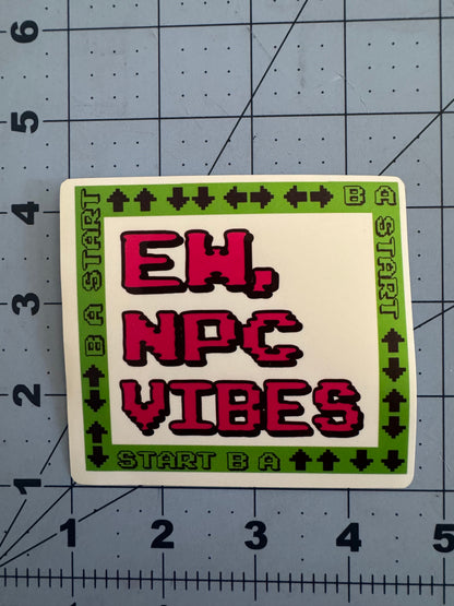 Two versions of a square sticker featuring pixelated text reading “Ew, NPC Vibes” in red on a white background. The border is bright green with black 8-bit arrow symbols and the words “START B A” repeated, referencing a classic video game controller pattern. One version has a standard white finish, the other has a holographic sheen. Stickers are shown on a blue grid cutting mat with inch measurements for scale.