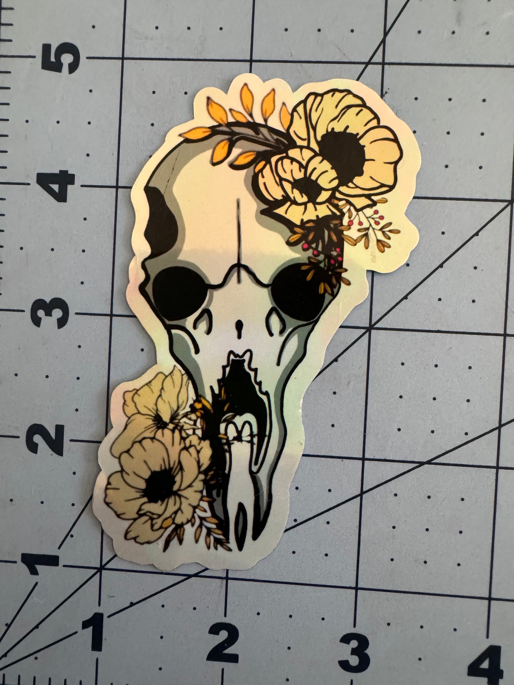 A holographic sticker featuring a stylized Fantasy steer skull adorned with yellow flowers and foliage. The skull has large, dark eye sockets and is surrounded by soft, natural floral elements on both the top and bottom.