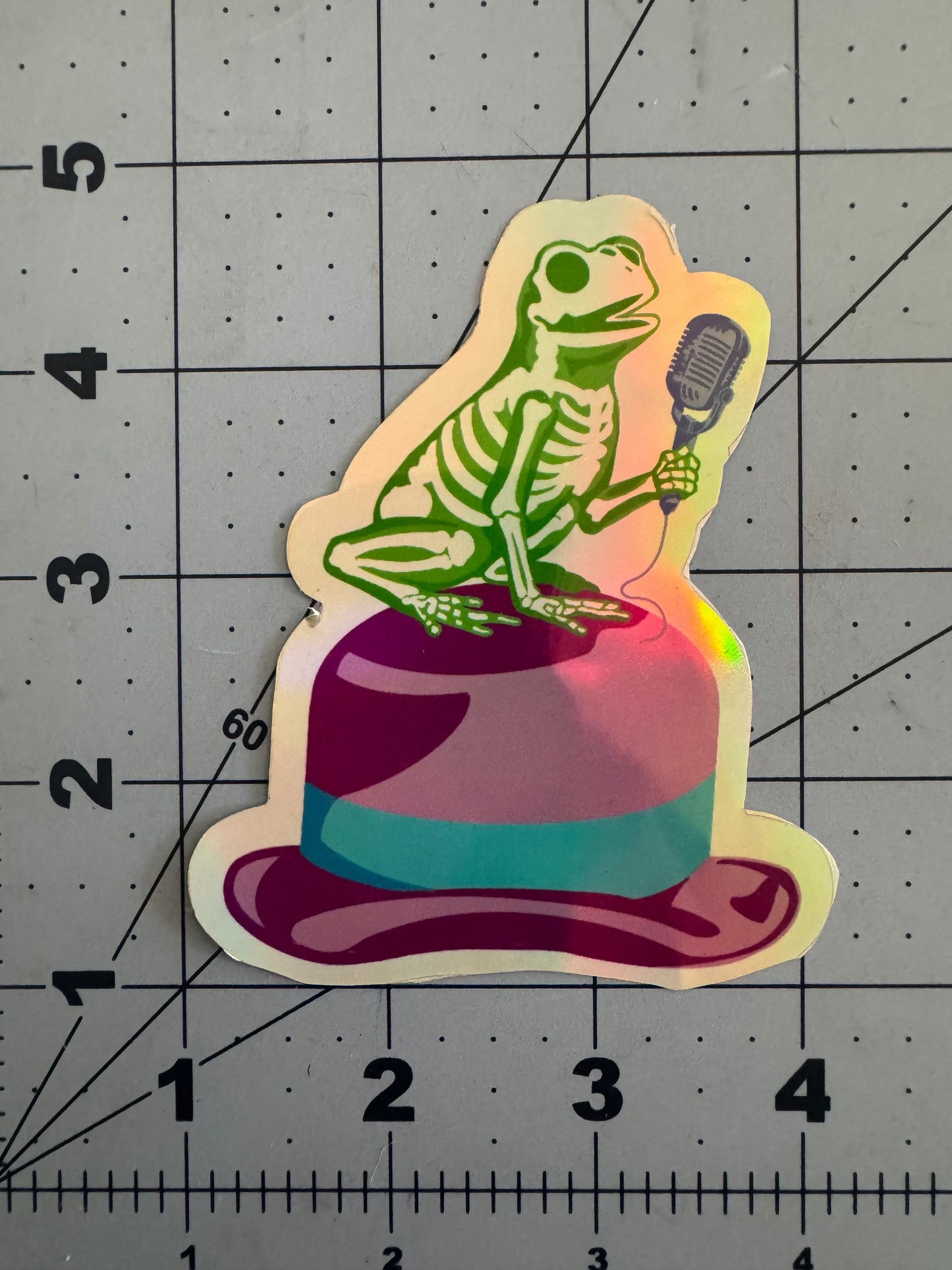 Holographic sticker of a green frog skeleton holding a vintage microphone while perched dramatically atop a pink and purple fedora with a teal band.
