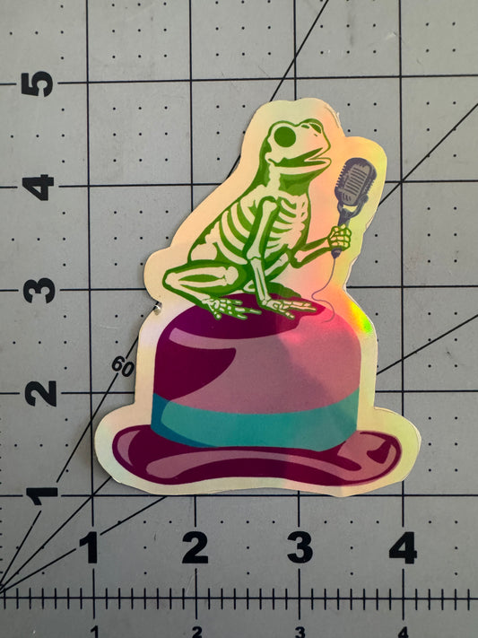 Holographic sticker of a green frog skeleton holding a vintage microphone while perched dramatically atop a pink and purple fedora with a teal band.