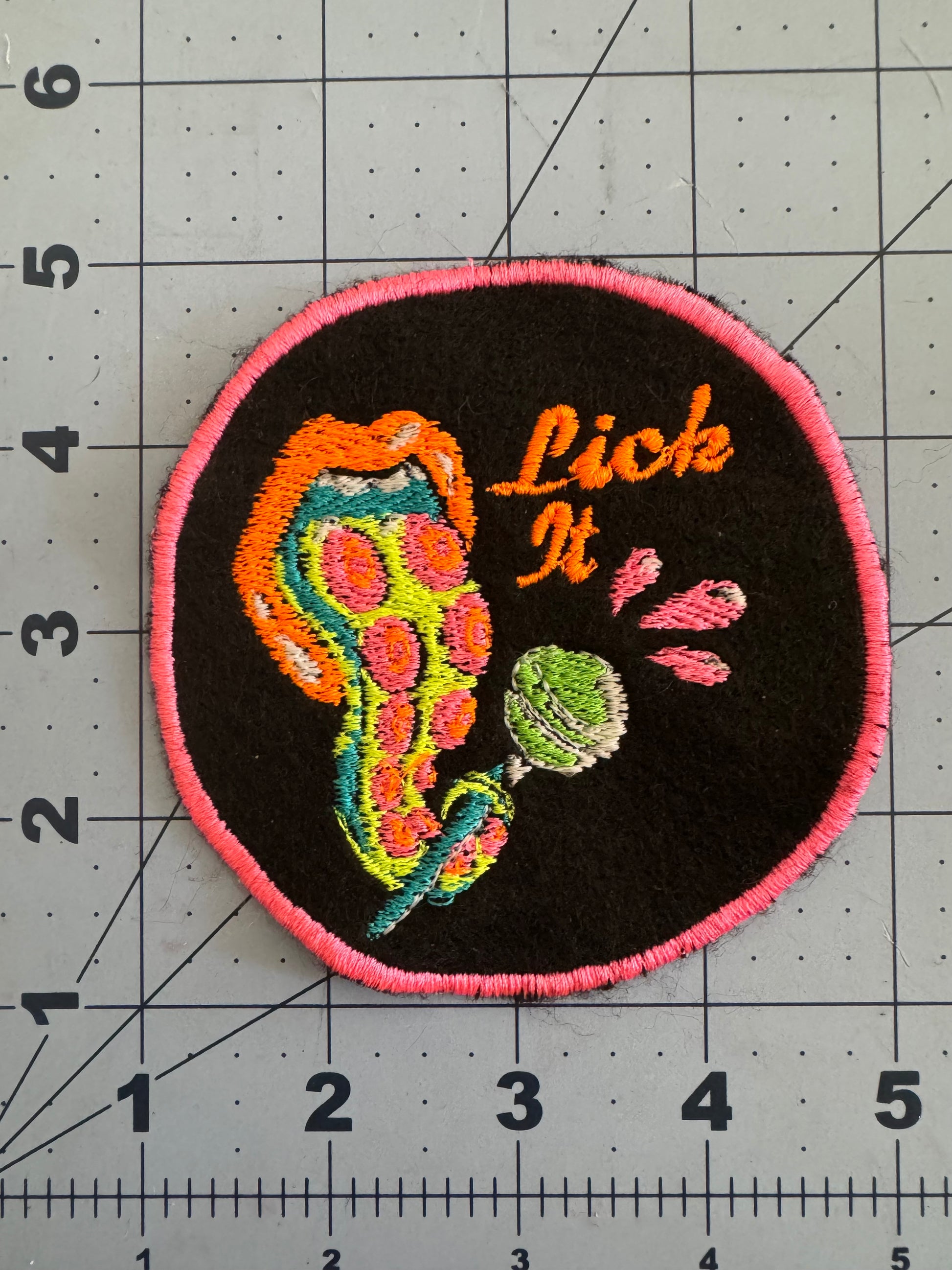 Embroidered patch featuring a psychedelic tentacle licking a green lollipop with the words “Lick It” stitched in orange. The patch has a bright pink border and glow-in-the-dark thread accents, set against a black felt background.
