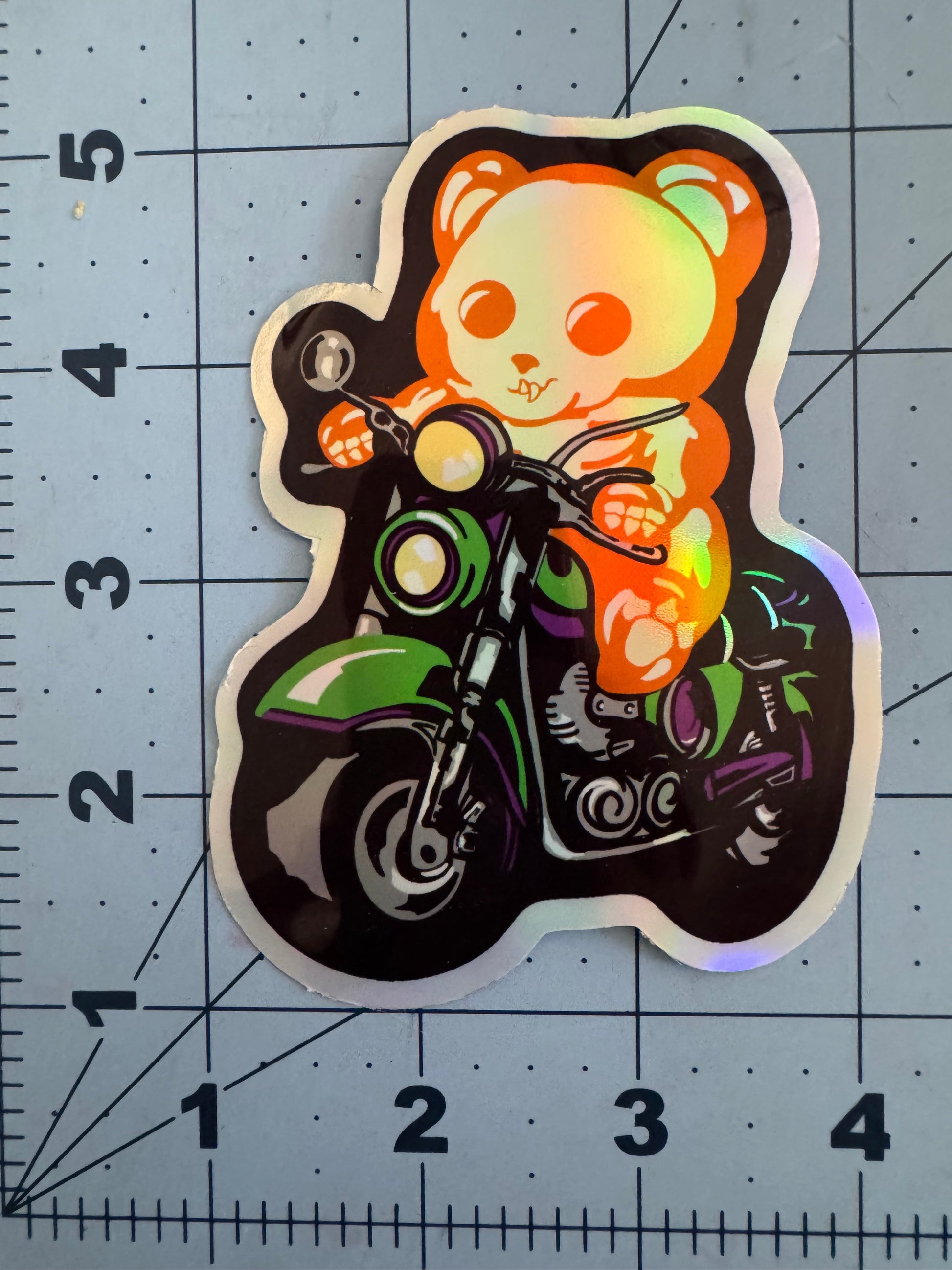 Holographic sticker of an orange skeletal gummy bear riding a green motorcycle, with bold black outlines and colorful accents. The sticker is placed on a cutting mat with a measurement grid, showing it is approximately 3 inches tall.