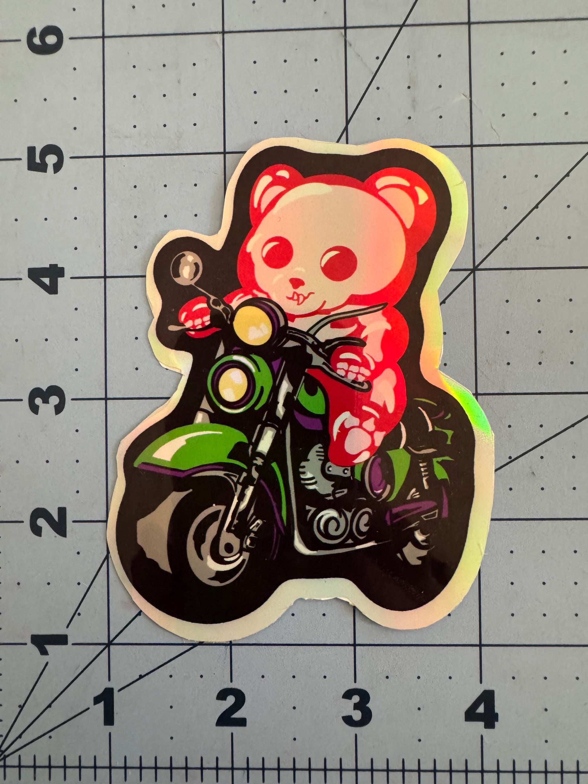 A holographic vinyl sticker featuring a red and white skeletal gummy bear riding a green and purple motorcycle. The bear grips the handlebars with a determined expression, surrounded by a glowing holographic finish that shifts colors under light. The sticker is placed on a blue grid background with inch measurements for scale.