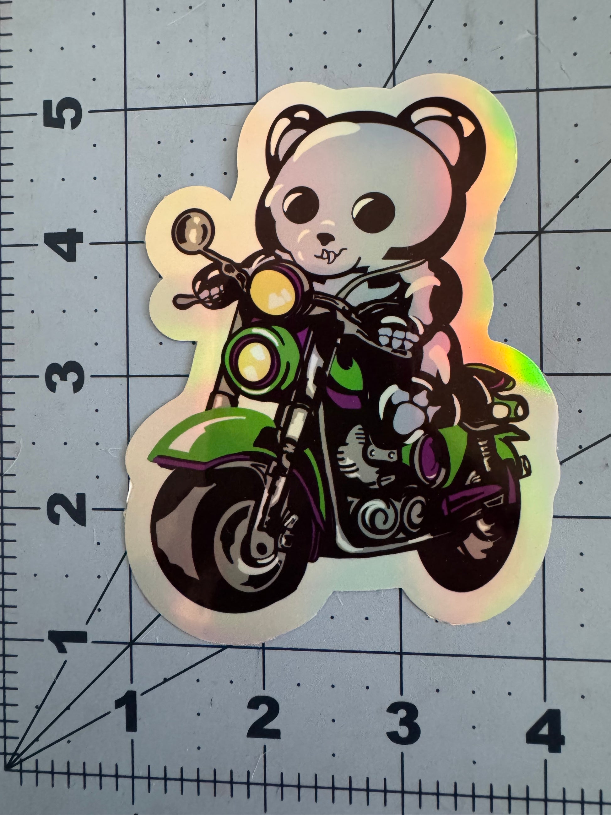 A holographic vinyl sticker featuring a black skeletal gummy bear riding a green and purple motorcycle. The bear grips the handlebars with a determined expression, surrounded by a glowing holographic finish that shifts colors under light. The sticker is placed on a blue grid background with inch measurements for scale.