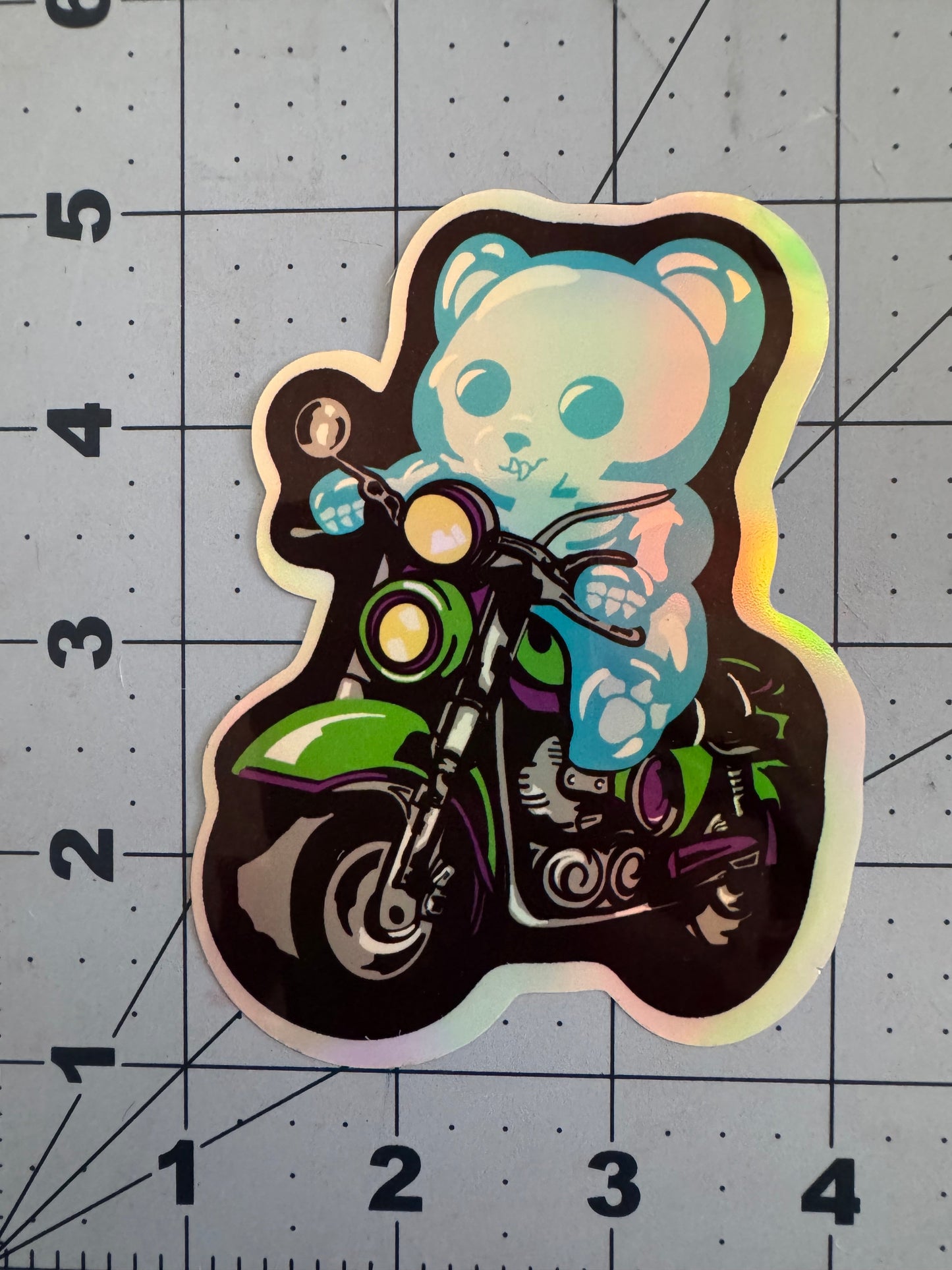 A holographic vinyl sticker featuring a blue skeletal gummy bear riding a green and purple motorcycle. The bear grips the handlebars with a determined expression, surrounded by a glowing holographic finish that shifts colors under light. The sticker is placed on a blue grid background with inch measurements for scale.