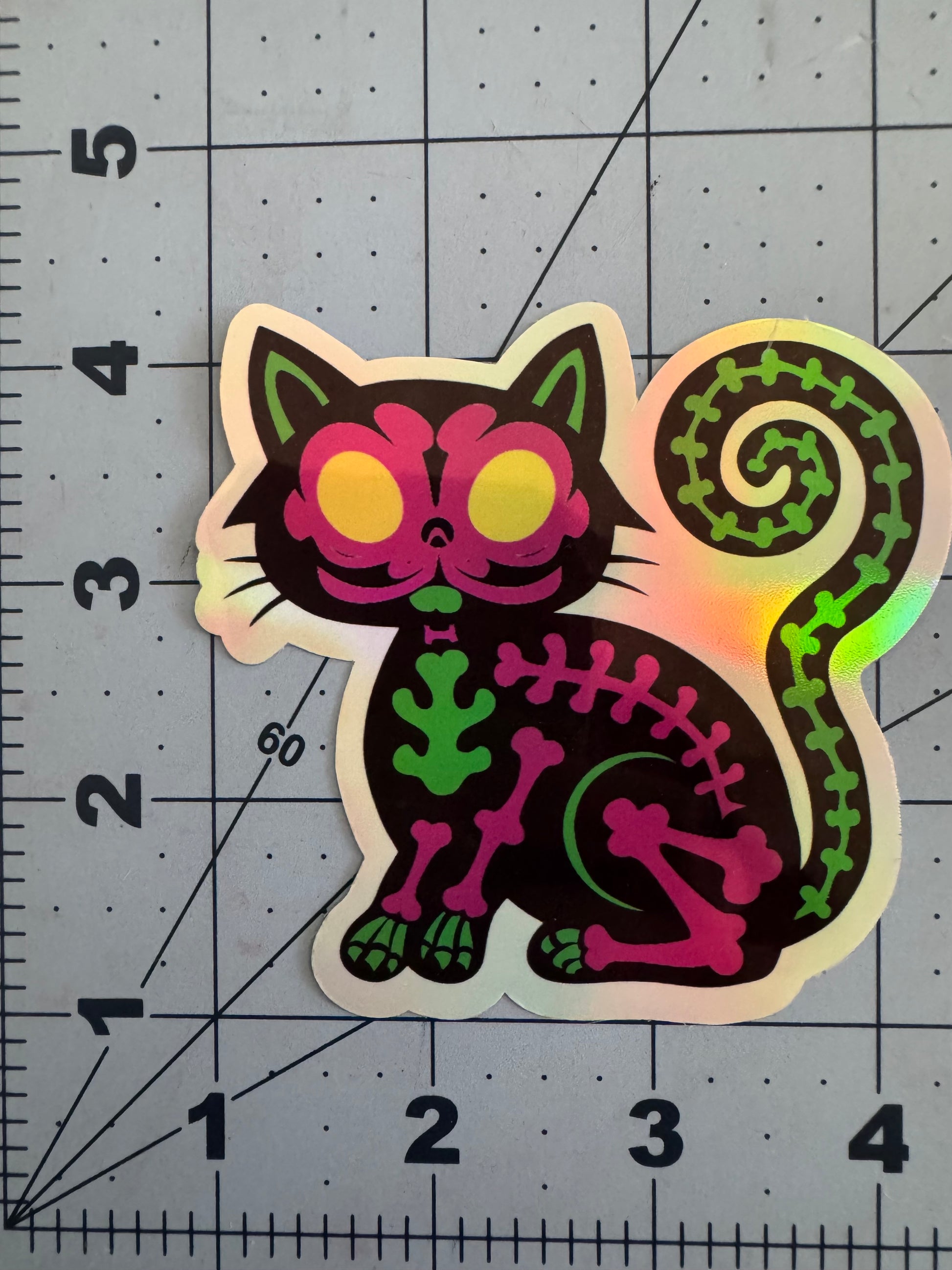 A holographic sticker of a black cat with neon skeleton bones in pink and green, yellow glowing eyes, and a spiraled tail made of vertebrae. The design is both spooky and playful, with a vibrant, cartoonish style.