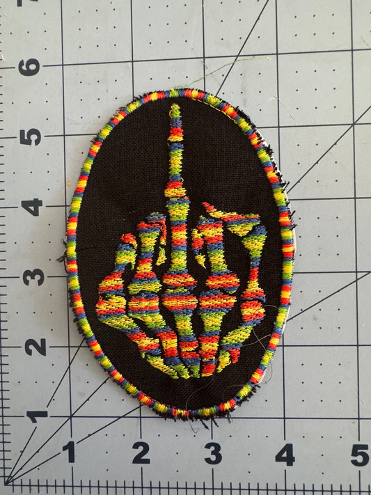An embroidered patch featuring a rainbow-striped skeleton hand giving the middle finger, set against a black background with an oval rainbow-thread border. The stitching is bold and colorful with visible texture, and the design measures approximately 3.5 by 5 inches.