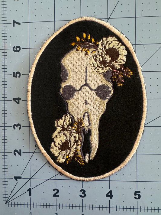 A beige and black embroidered patch featuring a skull adorned with sunflowers and small blooms, outlined with a light beige border.