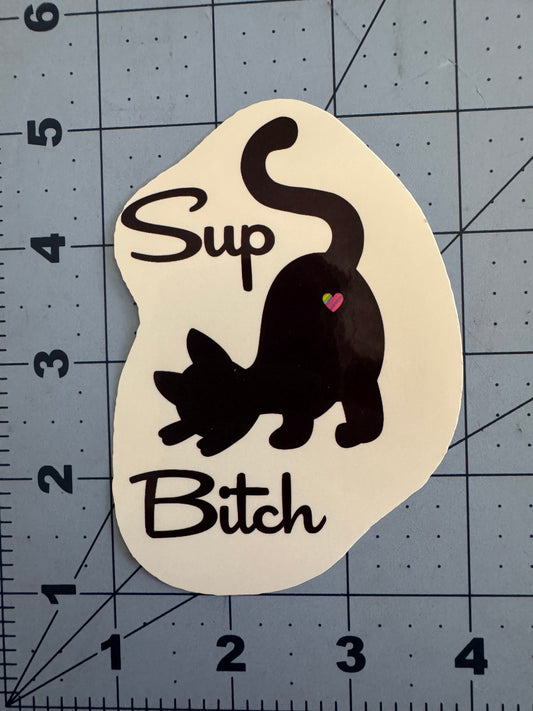 Sticker design featuring a black silhouette of a sassy cat with its tail up and a small rainbow heart on its rump. The text reads “Sup Bitch” in playful, handwritten-style font. Two versions are shown: one on a white background and one on a holographic finish.