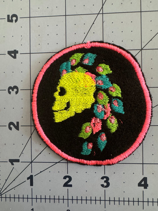 Two embroidered circular patches featuring side-profile skulls with bright mohawk hairstyles made of stylized neon flowers and leaves. One skull is yellow with pink and teal details and a hot pink border; the other is purple with multicolored flowers and a neon yellow border.