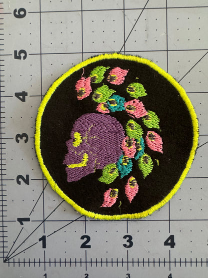 Two embroidered circular patches featuring side-profile skulls with bright mohawk hairstyles made of stylized neon flowers and leaves. One skull is yellow with pink and teal details and a hot pink border; the other is purple with multicolored flowers and a neon yellow border.