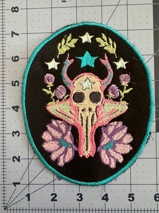 Embroidered patch featuring a pastel ram skull with floral antlers, surrounded by lavender flowers, thistle-like buds, and white and turquoise stars. Trimmed with a teal border on black fabric.