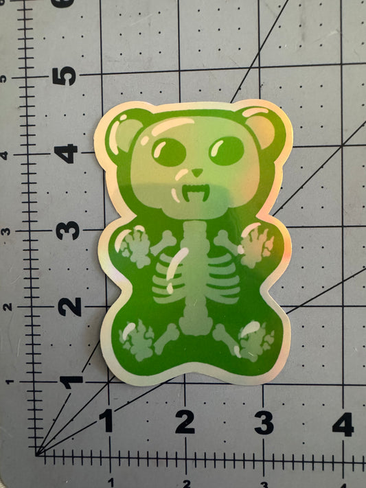 Two holographic vinyl stickers featuring cartoon-style gummy bears with fanged expressions and translucent bodies revealing skeletal structures inside; one is blue, the other green, both displayed on a measurement grid.