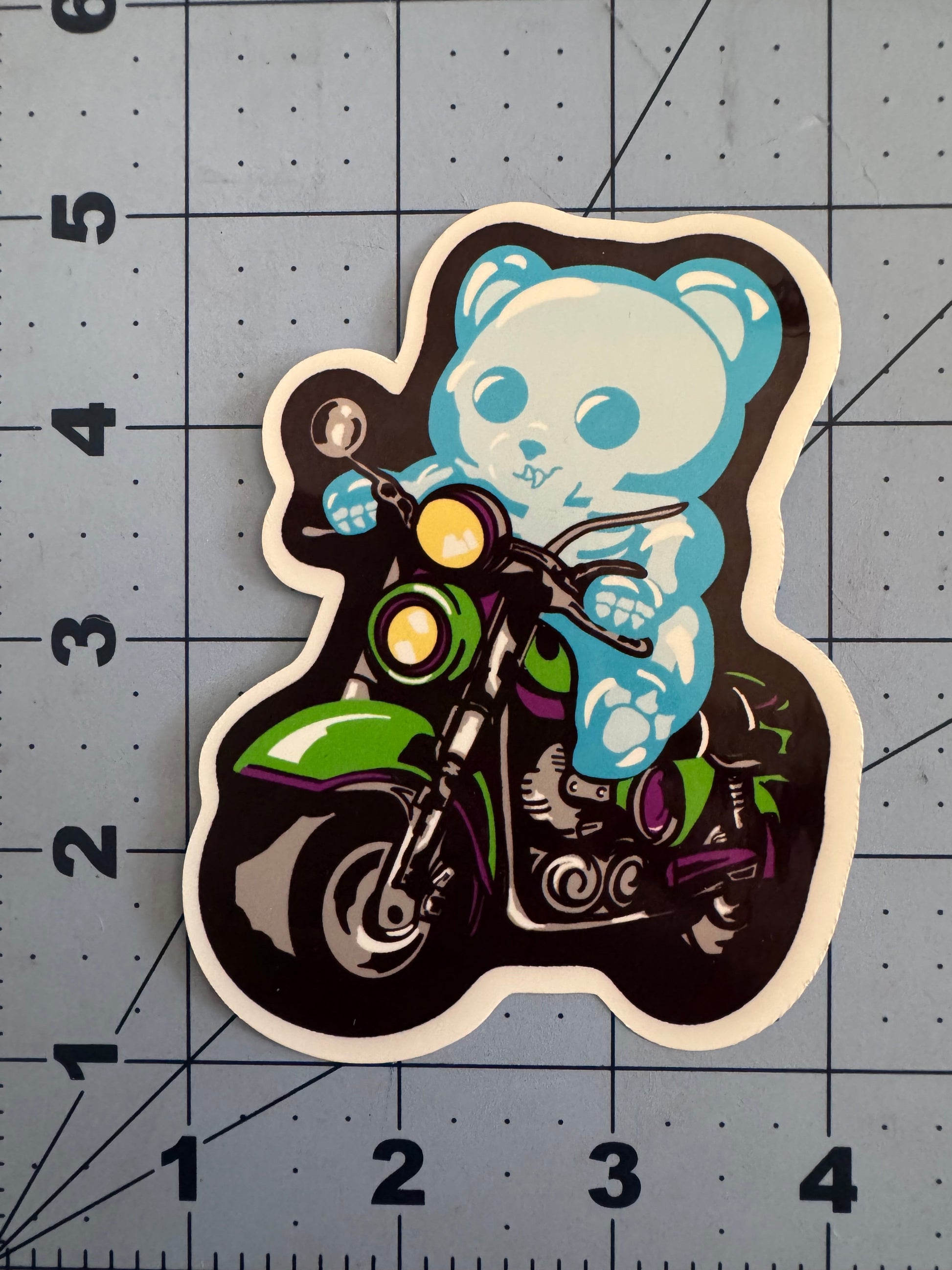 A classic white vinyl sticker featuring a blue skeletal gummy bear riding a green and purple motorcycle. The bear grips the handlebars with a determined expression. The sticker is placed on a blue grid background with inch measurements for scale.