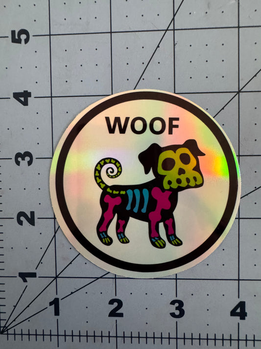 Holographic sticker featuring a cartoon skeleton dog with neon bones in blue, pink, and green on a black body. The word “WOOF” is printed above the dog in bold black letters, and the design is framed in a white circle with a black border.
